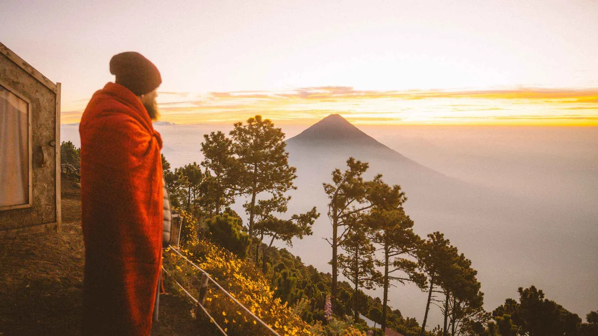 Hiker wrapped in a blanket at a cabin viewpoint watching sunrise over Volcan de Agua on a small-group Acatenango volcano tour, Guatemala